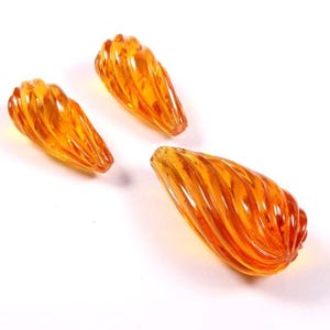 Puede incluir: Tres cuentas en forma de lágrima de color ámbar con un diseño en espiral. Las cuentas están hechas de un material translúcido y están dispuestas sobre un fondo blanco. Las cuentas tienen un acabado brillante y captan la luz.