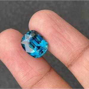 May include: A cushion-cut gemstone with a vibrant, deep blue color and a faceted surface that reflects light. The stone displays a clear, transparent quality with sharp geometric edges and a polished finish, showcasing a brilliant, saturated blue hue.