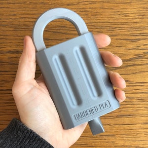 以下が含まれることがあります： ループハンドルとキーホールが付いたグレーの3Dプリントされたロック。ロックはアイスクリームの形をしており、側面に「HARDENED PLA」と印刷されています。