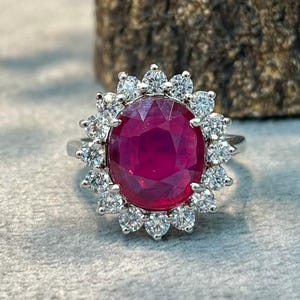 Könnte beinhalten: Ein silberfarbener Ring mit einem großen, oval geschliffenen, tiefroten Edelstein in der Mitte, der von einem Kranz aus kleinen, runden, klaren weißen Steinen umgeben ist. Der Ring hat eine polierte Metallschiene und ist frontal abgebildet.