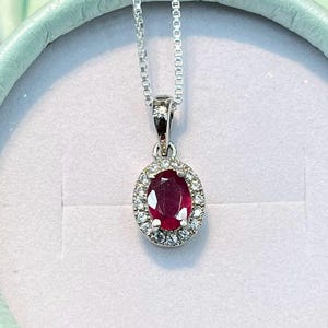 Op de afbeelding: Een zilveren ketting met een ovaalvormige rode robijn hanger omringd door een halo van kleine heldere steentjes. De hanger hangt aan een fijne zilveren ketting en wordt gepresenteerd in een rond, licht mintgroen sieradendoosje.