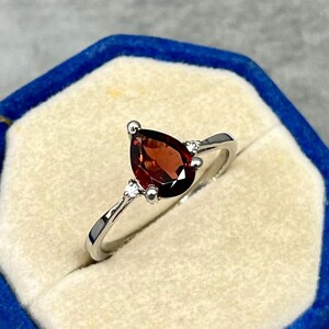 Op de afbeelding: Een zilverkleurige ring met een peervormige, dieprode granaat edelsteen als middelpunt, geflankeerd door twee kleine, heldere, rond geslepen accentstenen die aan weerszijden van de zetting in de ringband zijn geplaatst.