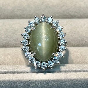 Puede incluir: Un anillo de tono plateado con una gran piedra preciosa ovalada de color verde oliva claro con efecto ojo de gato en el centro. La piedra central está rodeada por un halo de pequeñas piedras redondas, transparentes y brillantes en un engaste festoneado.