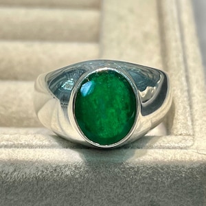Könnte beinhalten: Ein Silberring mit einem großen, runden, leuchtend grünen Stein in der Mitte. Der Ring hat ein glattes, poliertes Band und ein klassisches, elegantes Design. Der Edelstein hat einen tiefen, satten Grünton.