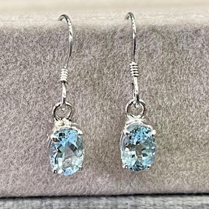 Può includere: Un paio di orecchini pendenti in argento con pietre preziose ovali azzurro chiaro. Gli orecchini hanno un design semplice con un gancio e un dettaglio in filo decorativo sopra la gemma.