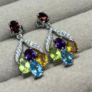 May include: Silver earrings with a cluster of gemstones in various colours. Each earring has a garnet stud, a teardrop design with clear stones, and a fan of oval-cut gems in purple, yellow, green, and blue.