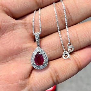 May include: A silver necklace with a teardrop-shaped pendant. The pendant features a vibrant red gemstone surrounded by a halo of smaller, clear gemstones. The chain is delicate and appears to be made of silver.