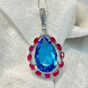 May include: A silver pendant necklace featuring a large, pear-shaped blue gemstone at the center, surrounded by a halo of small, oval-cut red gemstones. The pendant is suspended from a delicate silver chain, with small clear stones on the bail.