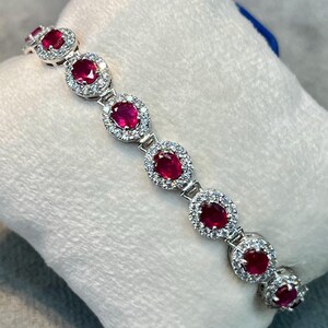 Pode incluir: Uma pulseira de elos em tom prateado com uma série de pedras preciosas ovais de um vermelho vibrante, cada uma rodeada por um halo de pequenas pedras claras e brilhantes. A pulseira é exibida sobre uma almofada branca acolchoada.