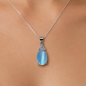 Puede incluir: Un collar de plata con un colgante en forma de lágrima azul claro. El colgante tiene un engaste de plata con un pequeño grupo de piedras azules en la parte superior. El collar se lleva alrededor del cuello.