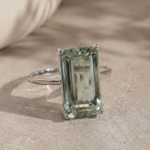 May include: A silver-toned ring featuring a large, rectangular, emerald-cut light green gemstone set in a four-prong setting. The band is thin and polished, showcasing a clear, faceted stone that reflects light across its surface.