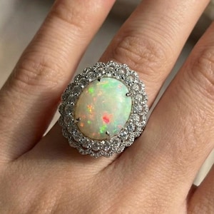 May include: An oval opal ring with a halo of small, clear gemstones. The opal displays a play of colour with flashes of green, red, and yellow. The ring is set in a silver or white gold band.