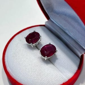 K&ouml;nnte beinhalten: Ohrstecker mit ovalen Rubinen in Silber, pr&auml;sentiert in einer roten Samt-Schmuckschatulle mit wei&szlig;em Innenraum. Die Ohrringe sind tiefrot und werden in einer runden, offenen Schatulle pr&auml;sentiert.