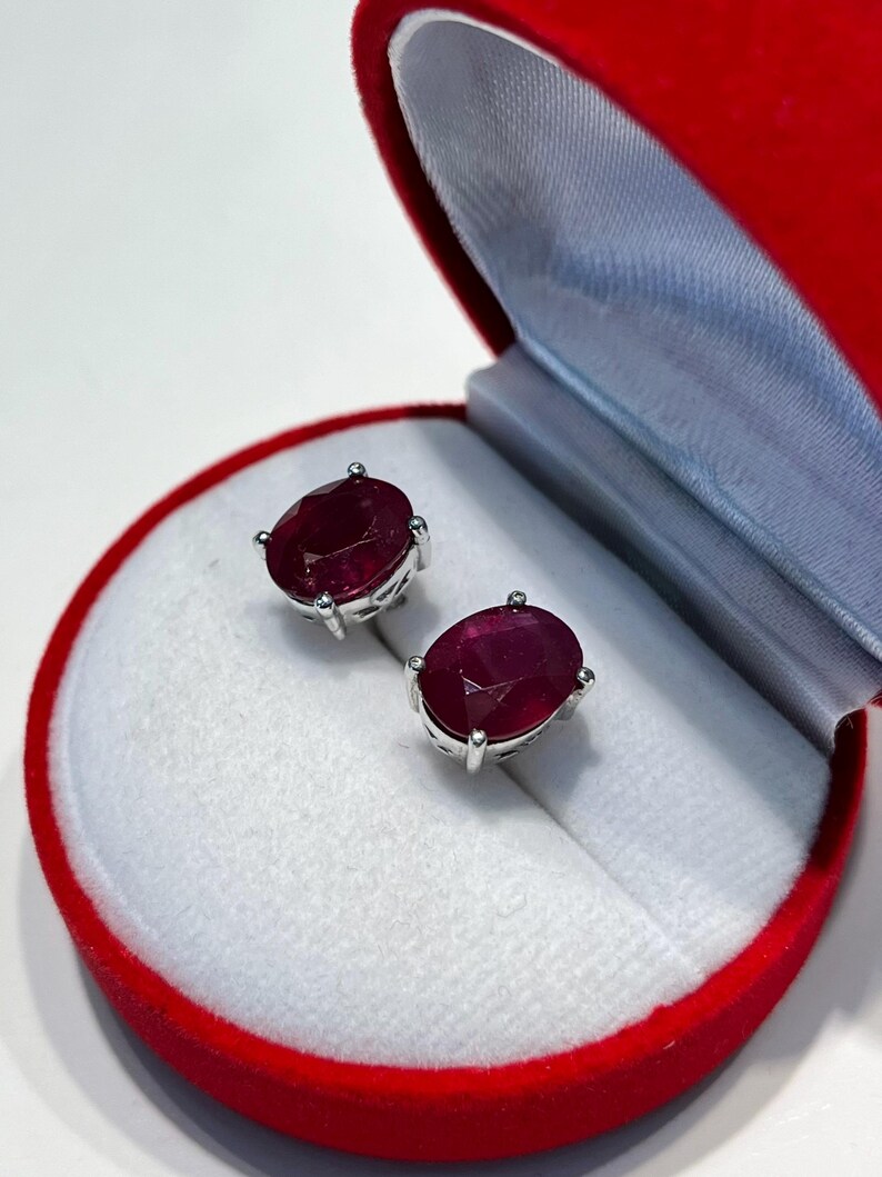 K&ouml;nnte beinhalten: Ohrstecker mit ovalen Rubinen in Silber, pr&auml;sentiert in einer roten Samt-Schmuckschatulle mit wei&szlig;em Innenraum. Die Ohrringe sind tiefrot und werden in einer runden, offenen Schatulle pr&auml;sentiert.