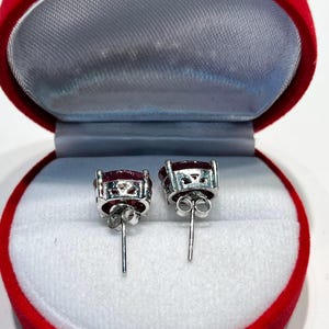 K&ouml;nnte beinhalten: Ein Paar Ohrstecker mit ovalen, tiefroten Edelsteinen, die in silberfarbenem Metall gefasst sind. Die Ohrringe werden in einer roten Samt-Schmuckschatulle mit grauem Innenraum pr&auml;sentiert.