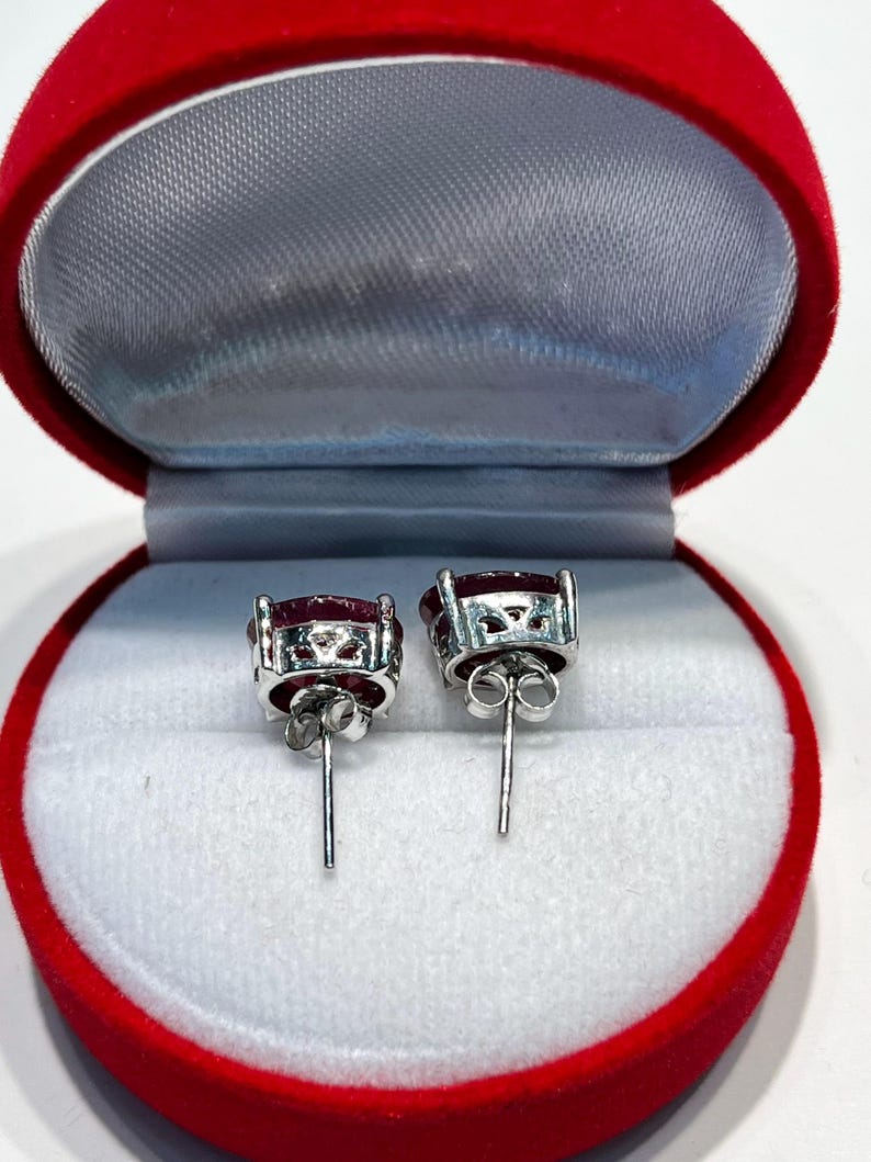 K&ouml;nnte beinhalten: Ein Paar Ohrstecker mit ovalen, tiefroten Edelsteinen, die in silberfarbenem Metall gefasst sind. Die Ohrringe werden in einer roten Samt-Schmuckschatulle mit grauem Innenraum pr&auml;sentiert.