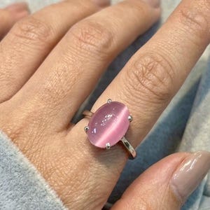 May include: A silver ring featuring an oval-shaped, light pink cat's eye stone set in a four-prong mounting. The smooth, polished band supports the cabochon gemstone, which displays a distinct vertical chatoyant band of light across its surface.