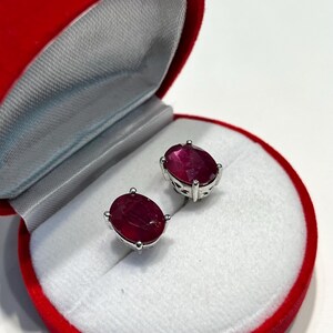 K&ouml;nnte beinhalten: Ohrringe mit oval geschliffenen Rubinen in Silberfassung, pr&auml;sentiert in einer roten Samt-Schmuckschatulle. Die tiefroten Rubine sind poliert. Die Schatulle ist ge&ouml;ffnet und zeigt ein wei&szlig;es Satin-Interieur.