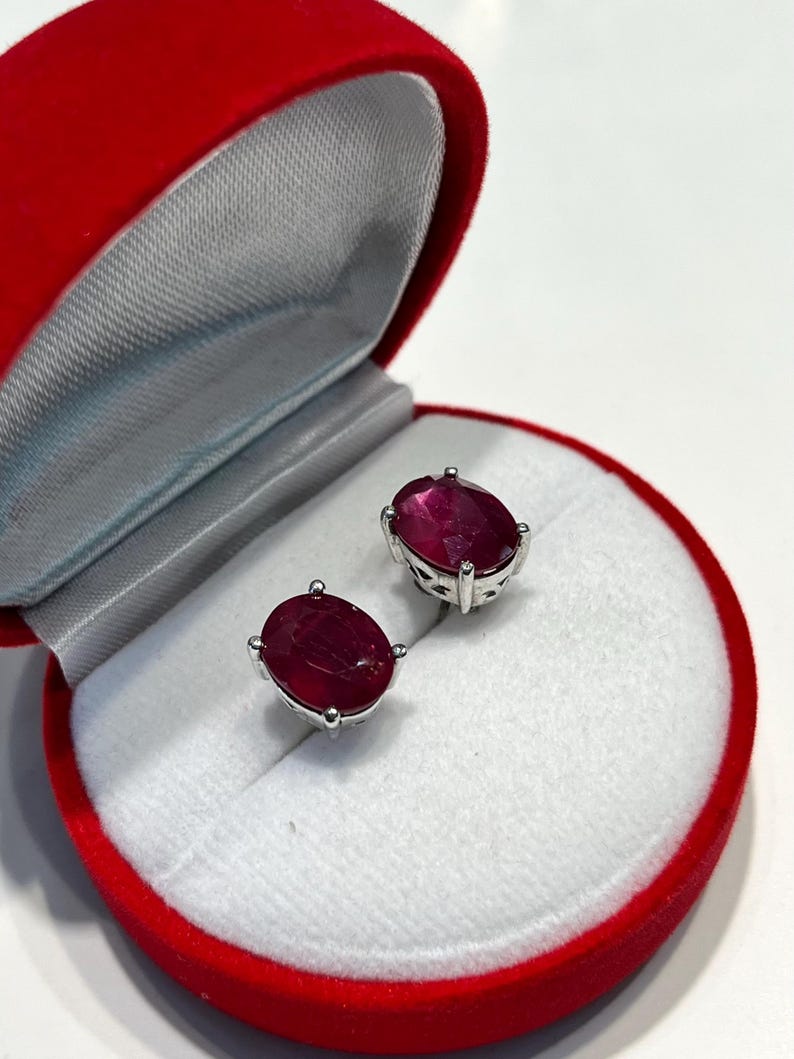 K&ouml;nnte beinhalten: Ohrringe mit oval geschliffenen Rubinen in Silberfassung, pr&auml;sentiert in einer roten Samt-Schmuckschatulle. Die tiefroten Rubine sind poliert. Die Schatulle ist ge&ouml;ffnet und zeigt ein wei&szlig;es Satin-Interieur.
