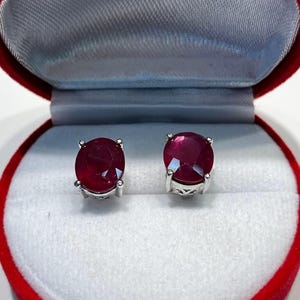 K&ouml;nnte beinhalten: Ein Paar Rubin-Ohrstecker, pr&auml;sentiert in einer roten Samt-Schmuckschatulle. Jeder Ohrring hat einen gro&szlig;en, runden, tiefroten Edelstein, der in einer silbernen Metalleinfassung gefasst ist. Die Schachtel hat ein wei&szlig;es Satin-Interieur.