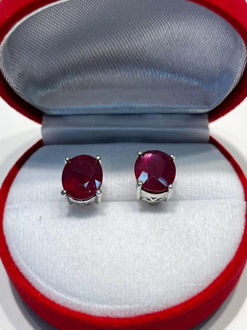 K&ouml;nnte beinhalten: Ein Paar Rubin-Ohrstecker, pr&auml;sentiert in einer roten Samt-Schmuckschatulle. Jeder Ohrring hat einen gro&szlig;en, runden, tiefroten Edelstein, der in einer silbernen Metalleinfassung gefasst ist. Die Schachtel hat ein wei&szlig;es Satin-Interieur.