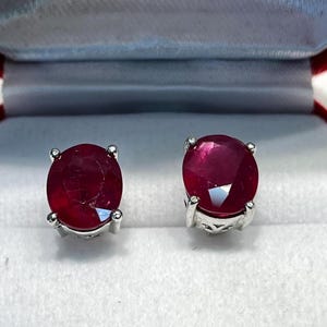 K&ouml;nnte beinhalten: Ein Paar Rubin-Ohrstecker. Jeder Ohrring hat einen runden, tiefroten Edelstein, der in einer silberfarbenen Metalleinfassung gefasst ist. Die Ohrringe werden in einer wei&szlig;en Schmuckschatulle mit rotem Innenraum pr&auml;sentiert.
