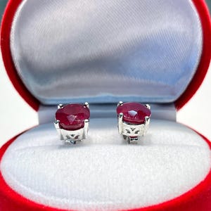 K&ouml;nnte beinhalten: Ein Paar Rubin-Ohrstecker, pr&auml;sentiert in einer offenen roten Schmuckschatulle mit wei&szlig;em Satin-Innenfutter. Die Ohrringe haben oval geschliffene, tiefrote Edelsteine, die in silberfarbenem Metall gefasst sind.