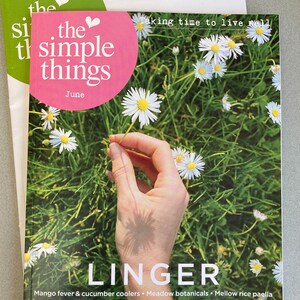 May include: The June issue of "The Simple Things" magazine features a hand holding a white daisy in a field of daisies. The magazine cover has the text "Taking time to live well" and "LINGER" in large white letters. The text below "LINGER" includes the words "Mango fever & cucumber coolers", "Meadow botanicals", "Mellow rice paella", "Sew-your-own cossie", "The magic of dawn & twilight", "Why we love a bench", "Swedish midsummer cake", "Gardens for rain or shine", and "Ley lines & holloways".