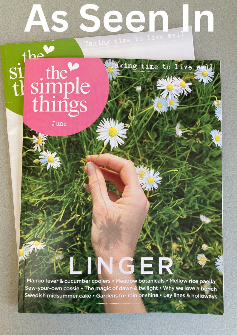 May include: The June issue of "The Simple Things" magazine features a hand holding a white daisy in a field of daisies. The magazine cover has the text "Taking time to live well" and "LINGER" in large white letters. The text below "LINGER" includes the words "Mango fever & cucumber coolers", "Meadow botanicals", "Mellow rice paella", "Sew-your-own cossie", "The magic of dawn & twilight", "Why we love a bench", "Swedish midsummer cake", "Gardens for rain or shine", and "Ley lines & holloways".