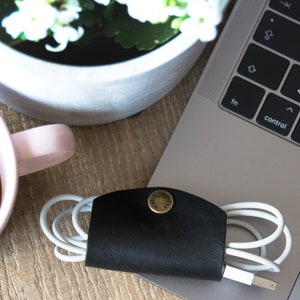 Könnte beinhalten: Ein schwarzer Leder-Kabelorganizer mit einem Messingknopf. Der Organizer hält ein weißes Ladekabel. Der Organizer befindet sich auf einer Holzoberfläche neben einem Laptop und einer Tasse Kaffee.