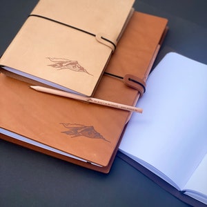 Op de afbeelding: Twee bruine lederen dagboeken met een bergontwerp en een wit dagboek met een zwarte lederen omslag. Een potlood ligt op de dagboeken.
