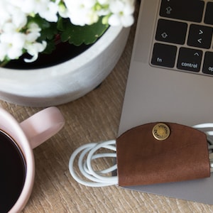 Könnte beinhalten: Ein brauner Leder-Kabelorganizer mit einem goldenen Metallknopf, auf dem "Unwind" steht. Der Organizer hält ein weißes Ladekabel.