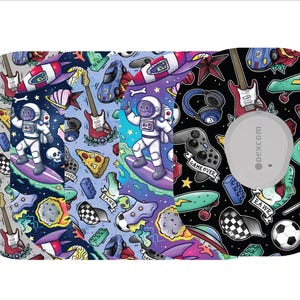 May include: A Dexcom G6 sensor patch with a colorful pattern featuring an astronaut surfing, guitars, spaceships, pizza slices, and skateboards. The design includes the text "GAME OVER" and "EAWZ" on a gray, blue, and black background.