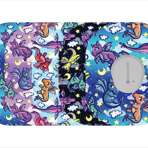 May include: A patterned medical device cover featuring colorful cartoon dragons in various colors against a night sky background with moons and stars. The cover has a gray circular device attached with the text "dexcom".