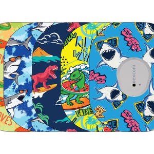May include: A collection of five round Dexcom G6 sensor covers with colorful designs. The covers feature illustrations of dinosaurs surfing, sharks with sunglasses, and tropical themes. One cover has a gray sensor attached.