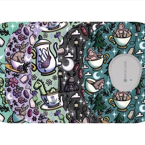 May include: A patterned medical device cover featuring whimsical illustrations. The design includes aliens, rabbits, dinosaurs, and Bigfoot characters, each in teacups, along with crystals, moons, and teapots. The cover has a gray circular component with the text "dexcom".