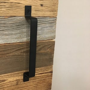 Puede incluir: Pomo de puerta de metal negro con forma rectangular y bordes redondeados. El pomo está montado en una puerta de madera con un acabado rústico.