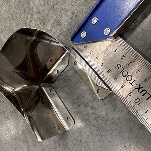 May include: A stainless steel corner bracket with a rounded edge, measured with a blue and silver ruler marked "LUX-TOOLS".