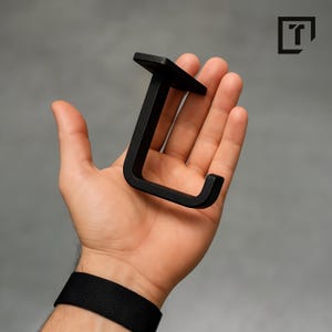 Può includere: Un gancio nero opaco a forma di J, con una parte superiore quadrata e una parte inferiore curva. Il gancio è tenuto nella mano di una persona su uno sfondo grigio. Il gancio sembra essere in metallo ed è progettato per appendere oggetti.