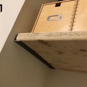 Könnte beinhalten: Ein Holzregal mit natürlicher Oberfläche ist mit schwarzen Metallkonsolen an einer Wand montiert. Das Regal wird von zwei Holzkisten mit Metall-Schubladengriffen getragen.