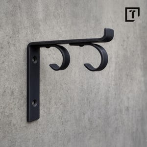 Può includere: Staffa da parete in metallo nero con due ganci curvi e una parte superiore orizzontale. La staffa è montata su un muro di cemento grigio. Il design include un supporto verticale con due fori per viti.