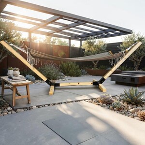 May include: A beige fabric hammock with white fringe trim suspended on a light-colored wooden frame with black metal joints. The hammock is positioned outdoors on a patio with a wooden bench nearby, featuring a clean, structural design.