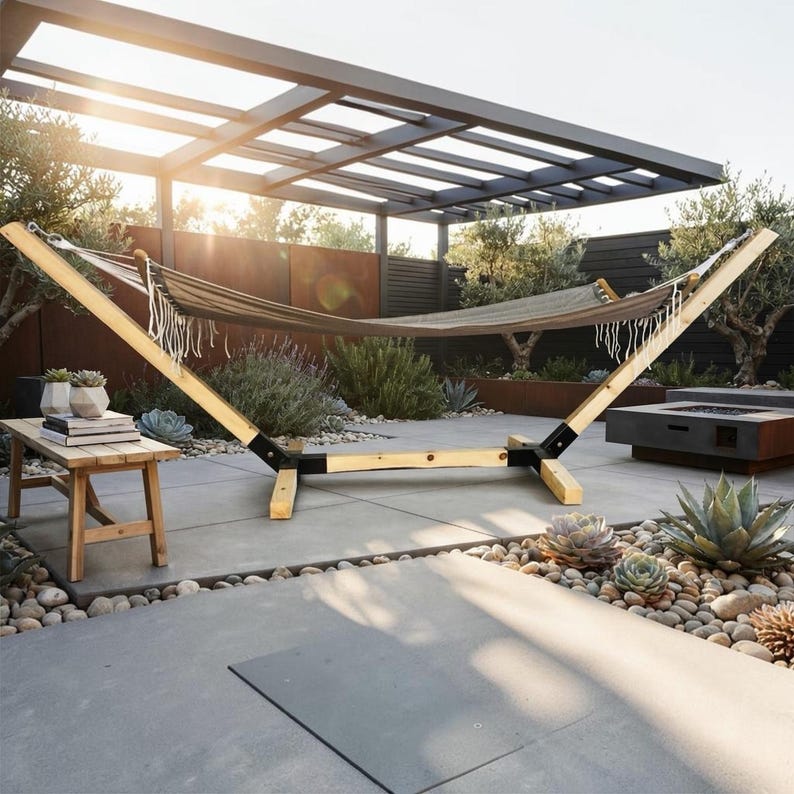May include: A beige fabric hammock with white fringe trim suspended on a light-colored wooden frame with black metal joints. The hammock is positioned outdoors on a patio with a wooden bench nearby, featuring a clean, structural design.