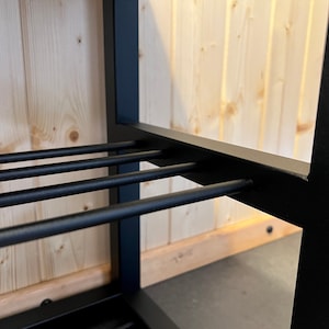 May include: Close-up of a black metal shelf with horizontal bars. The shelf is set against a light wood-panelled wall. The metal frame has a square profile, and the bars are evenly spaced.