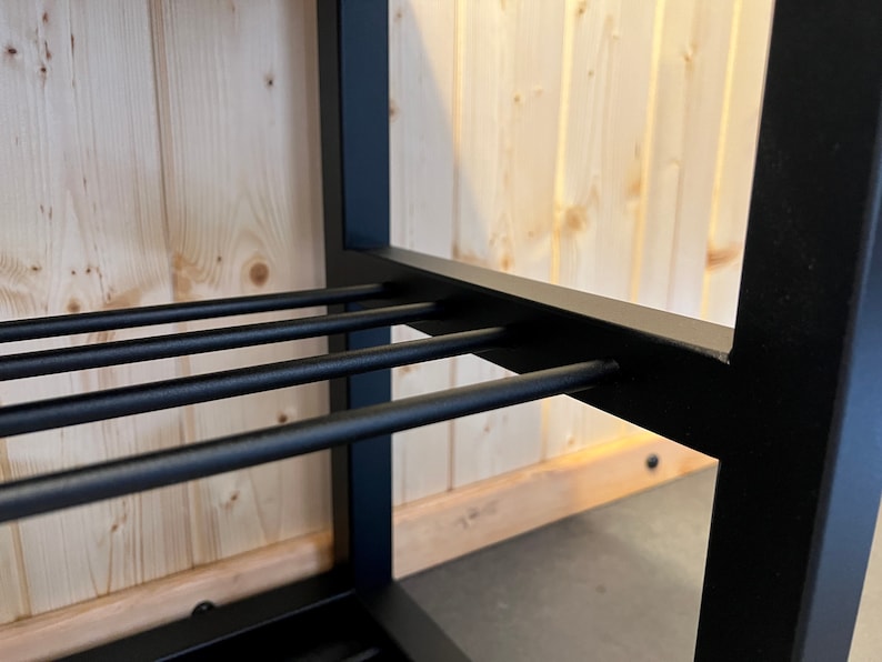 May include: Close-up of a black metal shelf with horizontal bars. The shelf is set against a light wood-panelled wall. The metal frame has a square profile, and the bars are evenly spaced.