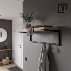 Peut inclure: Étagère murale moderne avec cadre en métal noir et étagère en bois clair. L'étagère comprend un porte-manteau intégré avec plusieurs crochets. Un pull gris est suspendu à l'un des crochets, et des clés à un autre. Une plante et une écharpe sont sur l'étagère.