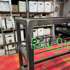 May include: A raw steel shelf unit with a rectangular frame and horizontal bars. The text "Raw Steel" is written in green. The unit is placed on a red and orange patterned surface.