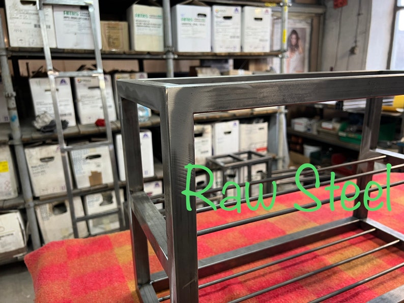 May include: A raw steel shelf unit with a rectangular frame and horizontal bars. The text "Raw Steel" is written in green. The unit is placed on a red and orange patterned surface.