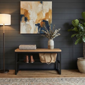 May include: An interior scene with a wooden bench with a black metal frame. The bench holds brown leather boots, a woven tote bag, and a folded blanket. An abstract painting hangs above, and a floor lamp and potted plant complete the decor.