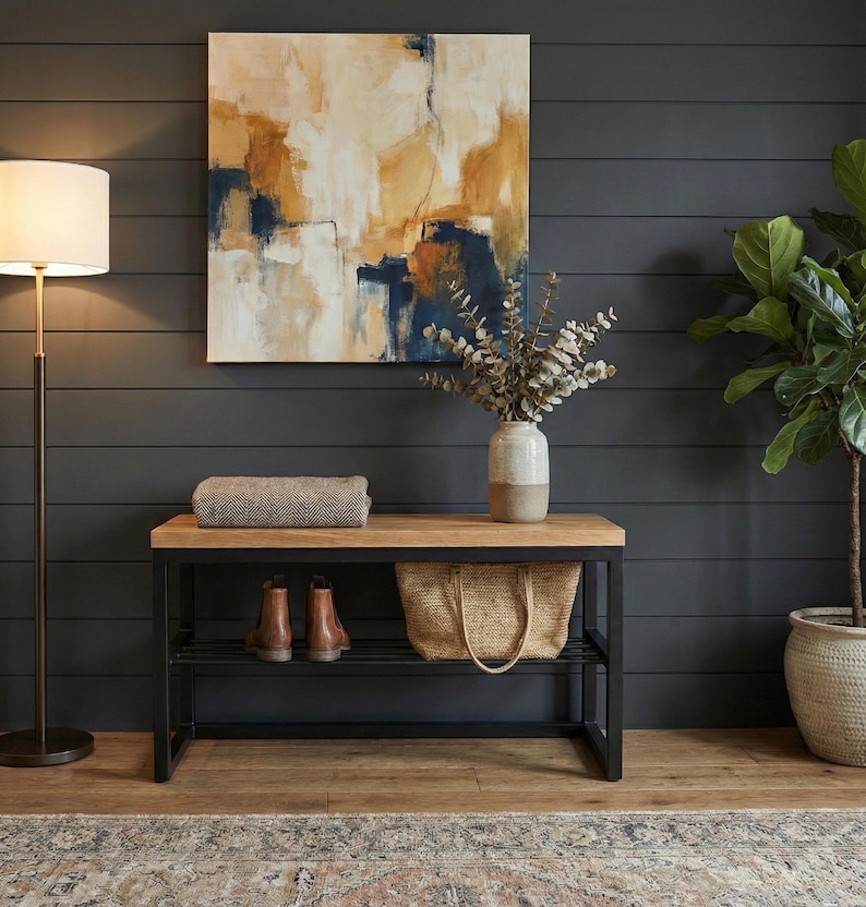 May include: An interior scene with a wooden bench with a black metal frame. The bench holds brown leather boots, a woven tote bag, and a folded blanket. An abstract painting hangs above, and a floor lamp and potted plant complete the decor.